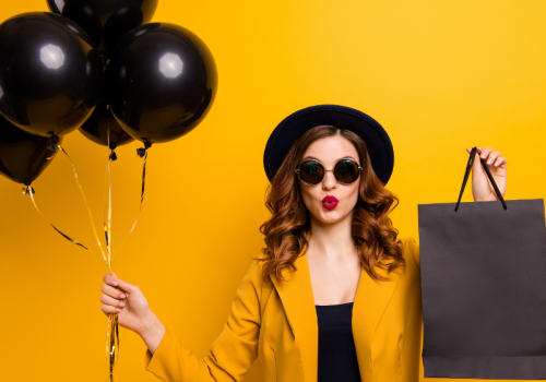 Eine Frau mit Sonnenbrille und Hut hält schwarze Luftballons und eine schwarze Einkaufstüte in den Händen - Titelbild zu "Der ultimative Black Friday-Survival Guide" / Foto von © deagreez via Adobe Stock