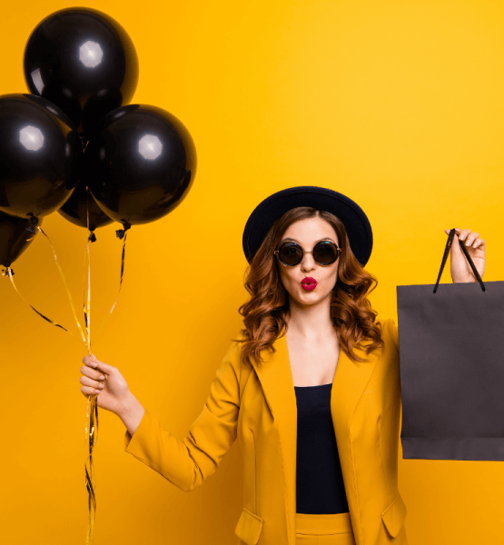 Eine Frau mit Sonnenbrille und Hut hält schwarze Luftballons und eine schwarze Einkaufstüte in den Händen - Titelbild zu "Der ultimative Black Friday-Survival Guide" / Foto von © deagreez via Adobe Stock