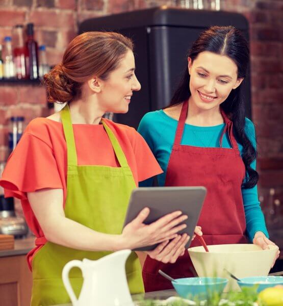 Fotografie von zwei Frauen, die an einem online Kochkurs teilnehmen. Eine der Frauen hält ein Tablet in den Händen und zeigt der anderen Frau, was sie auf dem Bildschirm erkennen kann.