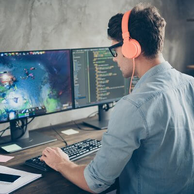 Wie erstelle ich ein eigenes Spiel? Mann sitzt vor zwei Monitoren. Rechts ist Code zu sehen Links ein Spiel. Er trägt Kopfhörer.