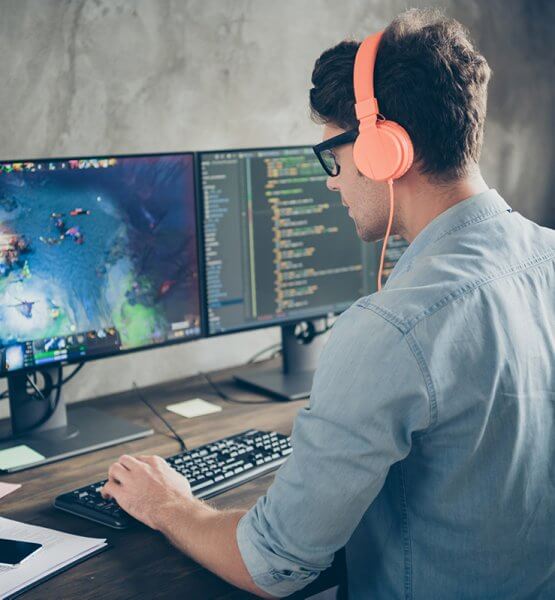 Wie erstelle ich ein eigenes Spiel? Mann sitzt vor zwei Monitoren. Rechts ist Code zu sehen Links ein Spiel. Er trägt Kopfhörer.