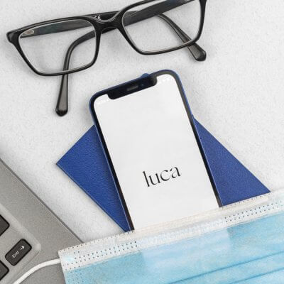 Smartphone hat die luca App geöffnet, über dem Smartphone liegt eine Brille, darunter eine Atemschutzmaske