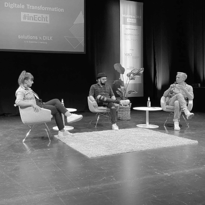 Live Aufzeichnung Podcast Tech und Trara mit Moritz und Marco Ilic und Sandra Müllrick auf einer Bühne. Alle sitzen zentral und auf Stühlen, Teppich auf dem Boden und Leinwand mit Bannern hinter ihnen