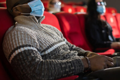 Mann mit Maske und 3D-Brille im Kino. Im Hintergrund zwei weitere Personen die auf ihren Plätzen sitzen.