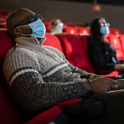 Mann mit Maske und 3D-Brille im Kino. Im Hintergrund zwei weitere Personen die auf ihren Plätzen sitzen.