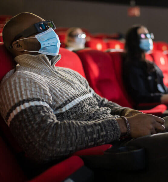 Mann mit Maske und 3D-Brille im Kino. Im Hintergrund zwei weitere Personen die auf ihren Plätzen sitzen.