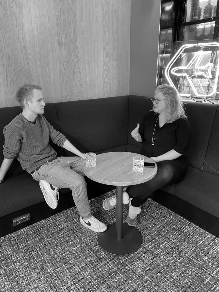 Julia Derndinger sitzt rechts am Bildrand, Moritz links, beide unterhalten sich und schauen sich dabei an. sie sitzen auf einer Bank mit Tisch und Getränken darauf