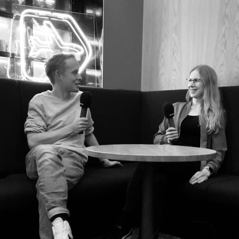 Hanna Huber und Moritz sitzen auf einer Bank, sie halten Mikrofone in den Händen, schauen sich an und lächeln freundlich