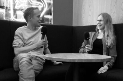 Moritz sitzt mit Hanna Huber auf einer Eckbank, er links, sie rechts, sie halten Mikrofone in der Hand und lächeln