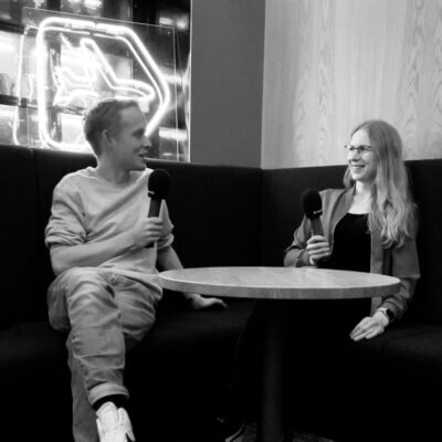 Moritz sitzt mit Hanna Huber auf einer Eckbank, er links, sie rechts, sie halten Mikrofone in der Hand und lächeln