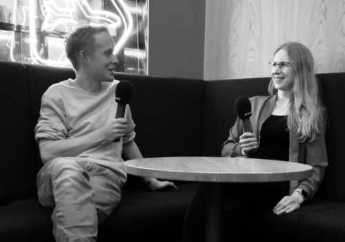 Moritz sitzt mit Hanna Huber auf einer Eckbank, er links, sie rechts, sie halten Mikrofone in der Hand und lächeln