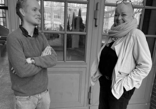 Moritz links, schaut in die Ferne mit verschränkten Armen, Sabine Fischer rechts schaut lachend in die Kamera, beide stehen vor dem Amplifier Berlin