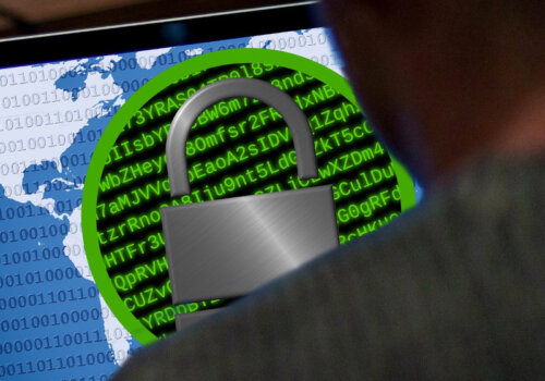 Mann sitzt vor einem PC. Der Bildschirm ist mit einsen und nullen beschrieben in der Mitte eine Art Meldung und ein Schloss, dass für Cybersicherheit steht.