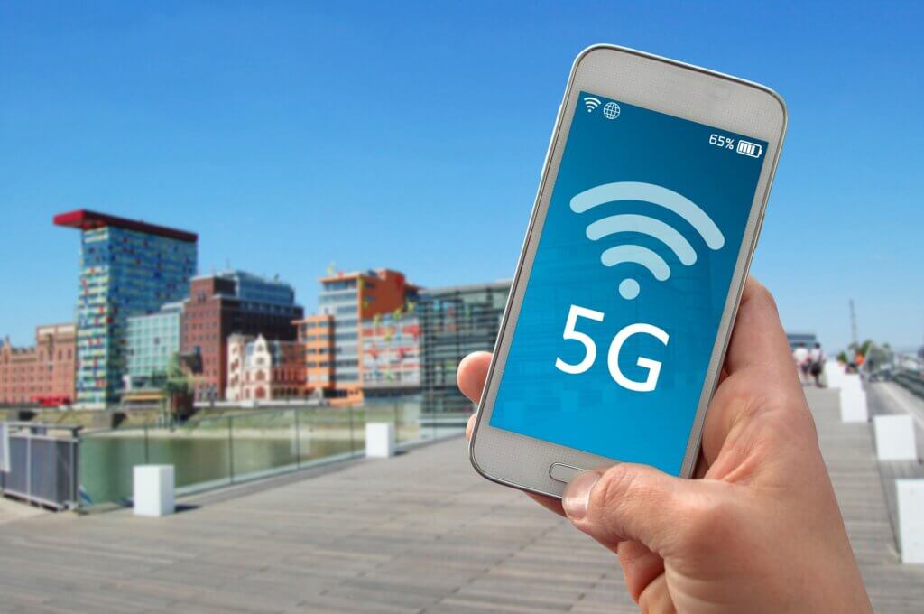 Ein Smartphone in der Hand, auf dem eine 5G-Verbindung ist. Im Hintergrund sieht man eine Stadt.