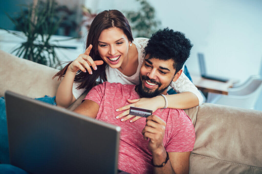 Ein Paar sitzt gemeinsam vorm Laptop und bestellt etwas in einem Onlineshop. Einer der beiden hält eine Kreditkarte in der Hand.
