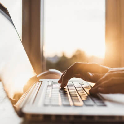 Eine Hand tippt auf einem Laptop, im Hintergrund scheint die Sonne der aufgehenden Sonne durch Fenster.