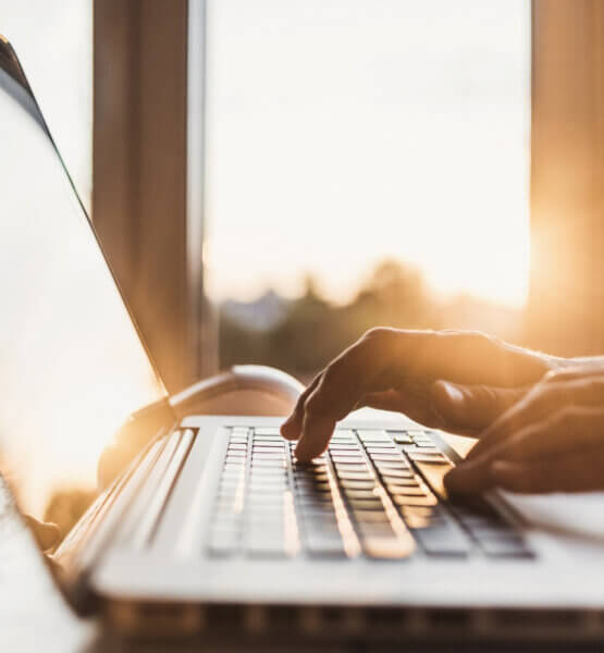 Eine Hand tippt auf einem Laptop, im Hintergrund scheint die Sonne der aufgehenden Sonne durch Fenster.