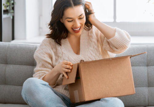 Eine Frau sitzt im Schneidersitz auf dem Sofa und schaut freudig in ein Paket, dass sie gerade geöffnet hat.