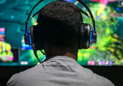 Man sieht einen jungen Mann von hinten, der vor einem Monitor sitzt. Er trägt ein Gaming-Headset mit leichter Beleuchtung.