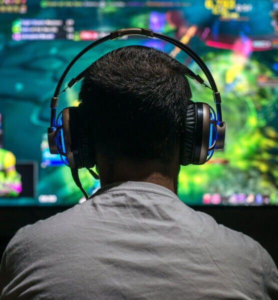 Man sieht einen jungen Mann von hinten, der vor einem Monitor sitzt. Er trägt ein Gaming-Headset mit leichter Beleuchtung.