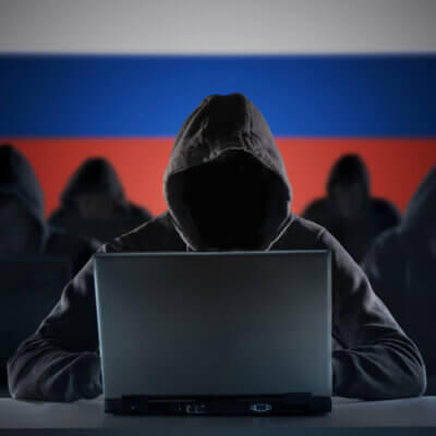 Eine Gruppe unkenntlicher Personen in Kapuzenhoodies arbeiten an Laptops. Der Hintergrund ist eine russische Flagge.