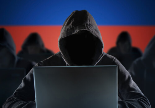 Eine Gruppe unkenntlicher Personen in Kapuzenhoodies arbeiten an Laptops. Der Hintergrund ist eine russische Flagge.