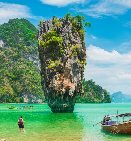 Reisender mit Selfiestick knietief im Meer in Thailand. Im Hintergrund atmenberaubende Berge. Handelt es sich um einen Travel-Vlogger?