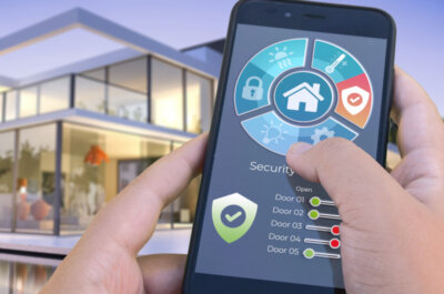 Eine Person steht mit dem Smartphone in der Hand vor einem Haus und kann damit das Smarthome, insbesondere die Sicherheit steuern.