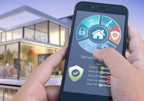 Eine Person steht mit dem Smartphone in der Hand vor einem Haus und kann damit das Smarthome, insbesondere die Sicherheit steuern.