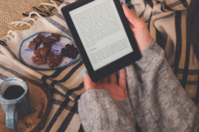 Beitragsbild die besten E-Book-Reader: Draufsicht einer Frau, die ein E-Book im E-Book-Reader mit einer Tasse Kaffee und Kakaokeksen liest. Gemütlicher Wintertag zu Hause und Hygge-Konzept. Der Text auf dem E-Book-Reader ist ein „Lorem Ipsum“