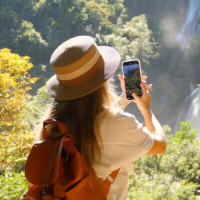 Eine Frau mit Hut und Rucksack steht in der unberührten Natur vor einem Wasserfall und hält den Moment mit ihrem Kamera-Handy fest.