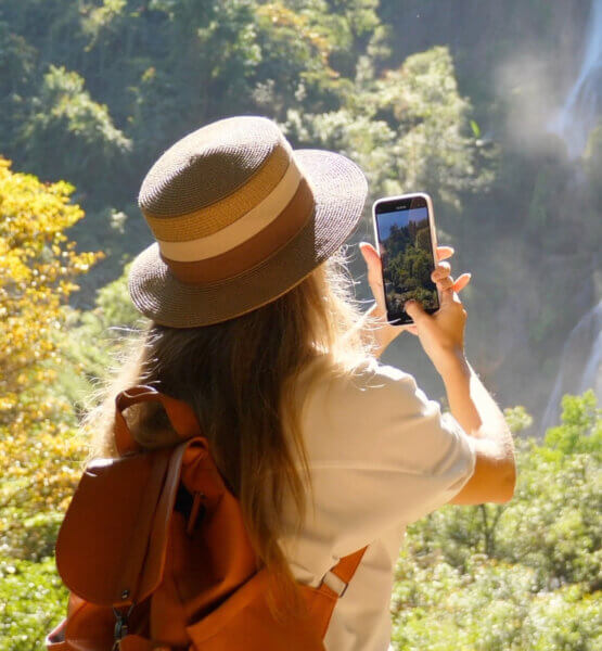 Eine Frau mit Hut und Rucksack steht in der unberührten Natur vor einem Wasserfall und hält den Moment mit ihrem Kamera-Handy fest.