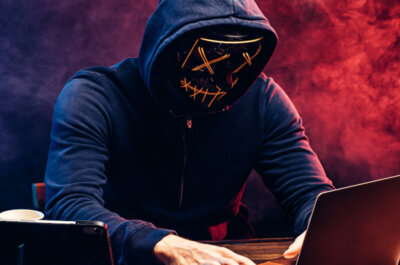 Hacker an einem Laptop, getarnt mit einer Maske mit aufgesticktem und etwas unheimlichen Smiley.
