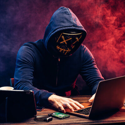 Hacker an einem Laptop, getarnt mit einer Maske mit aufgesticktem und etwas unheimlichen Smiley.