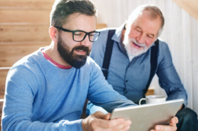 Ein Erwachsener und ein Senior sitzen zusammen auf der Treppe und schauen auf ein Tablet.