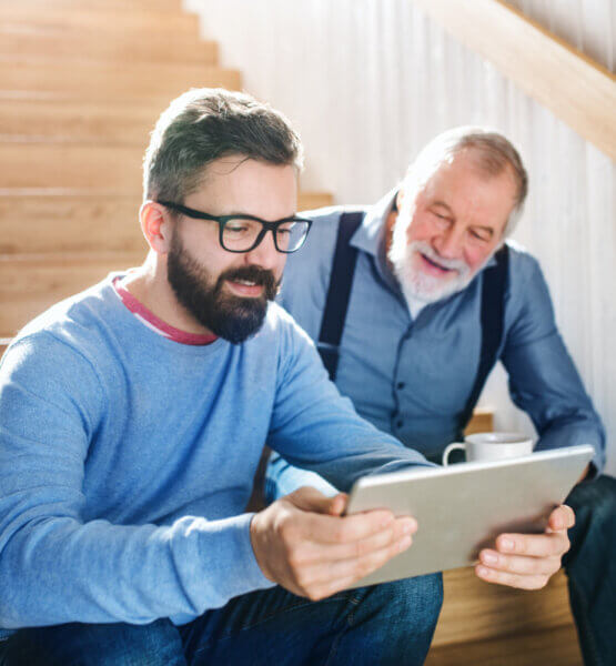 Ein Erwachsener und ein Senior sitzen zusammen auf der Treppe und schauen auf ein Tablet.