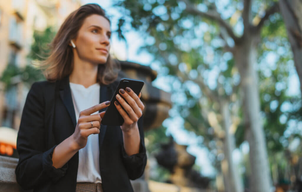 Eine Frau bewegt sich mit Smartphone durch eine grüne Stadt, der Blick dabei auch zufrieden auf die Umgebung gerichtet. 