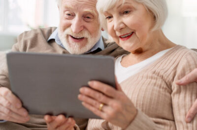 Zwei Senioren sitzen gutgelaunt auf dem Sofa und schauen auf ein Tablet, dass sie gemeinsam halten.