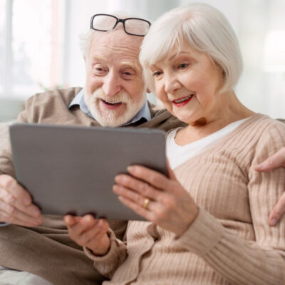 Zwei Senioren sitzen gutgelaunt auf dem Sofa und schauen auf ein Tablet, dass sie gemeinsam halten.