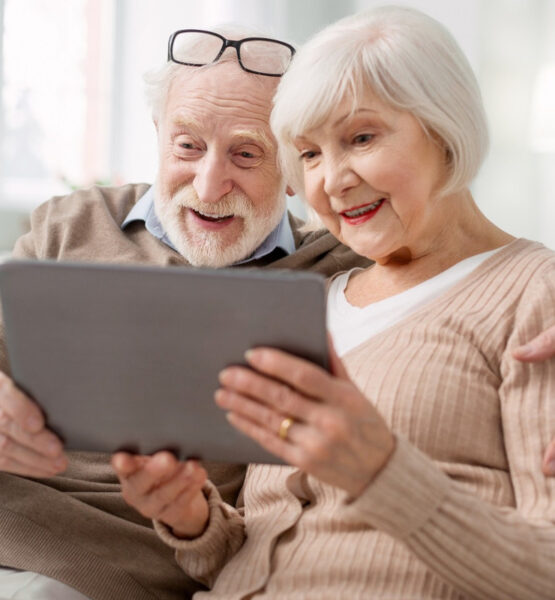 Zwei Senioren sitzen gutgelaunt auf dem Sofa und schauen auf ein Tablet, dass sie gemeinsam halten.