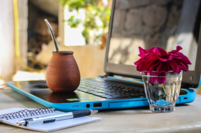 Ein blauer Laptop steht auf einem Außentisch mit einem Notizbuch und einer kleinen Blumenvase daneben.