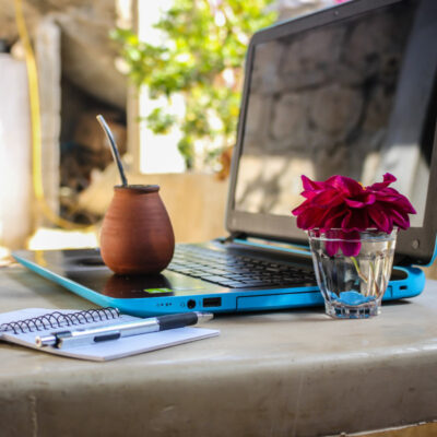 Ein blauer Laptop steht auf einem Außentisch mit einem Notizbuch und einer kleinen Blumenvase daneben.
