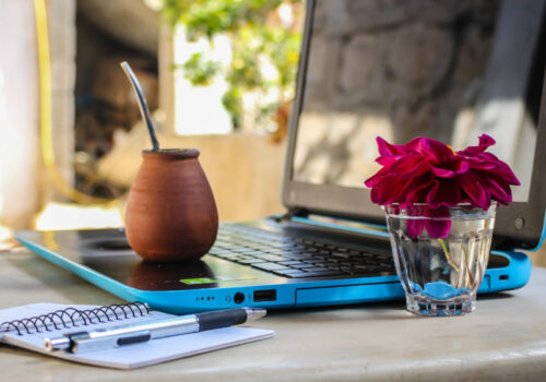 Ein blauer Laptop steht auf einem Außentisch mit einem Notizbuch und einer kleinen Blumenvase daneben.