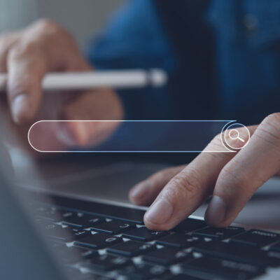 Eine Person tippt auf der Tastatur eines Laptops. In der Mitte des Bildes ist eine leere Suchmaske eingeblendet.