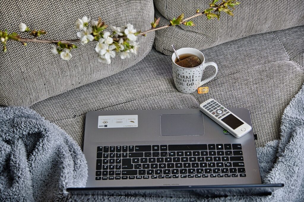 Laptop, Tee, Handy und Kuscheldecke auf einem grauen Sofa.