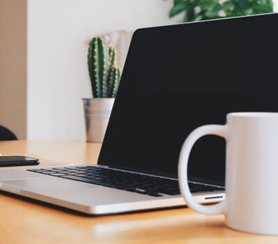 Ein Laptop zusammen mit einer Kaffeetasse und einem Smartphone auf einem Schreibtisch in einem hellen Raum.