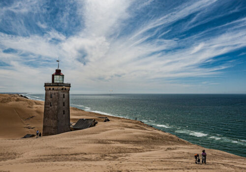 Ein schöner Sandstrand in Dänemark bei schönem Wetter mit einem Leuchtturm und nur vereinzelten Personen, die am Strand entlang wandern.