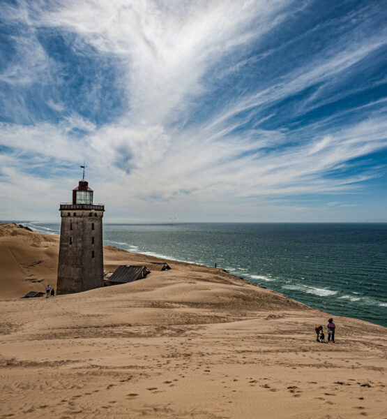 Ein schöner Sandstrand in Dänemark bei schönem Wetter mit einem Leuchtturm und nur vereinzelten Personen, die am Strand entlang wandern.