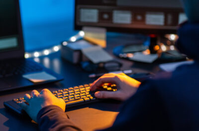 Person sitzt in einem dunklen Raum am PC und tippt auf der Tastatur.