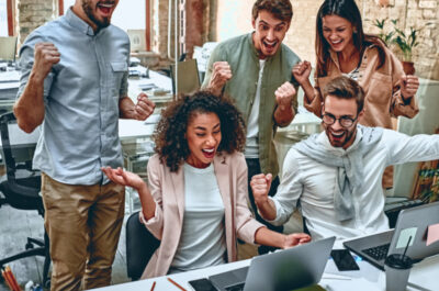 Mehrere Personen stehen und sitzen in einem Büro um einen Laptop und freuen sich über etwas auf dem Bildschirm.